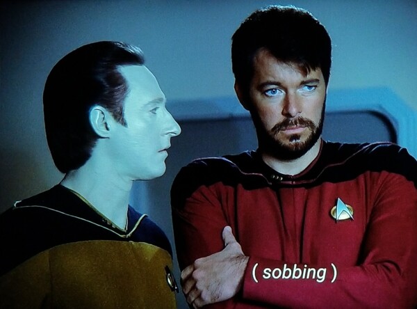 Early TNG scene. We're close in on Riker and Data both in uniform (Riker red, Data gold). Riker is arms crossed kind of holding himself and looking stumped at something in front of him. Data is standing very close (bad observance of the personal bubble rule) and looking directly at him from the side. Closed caption reads, "(sobbing)”