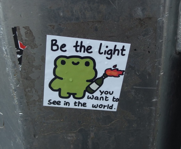 Sticker on a pole featuring a frog(?) carrying a Molotov and the words "Be the light you want to see in the world"