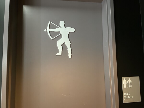 The door to a men’s bathroom with a silhouette of an archer, with short hair and pants